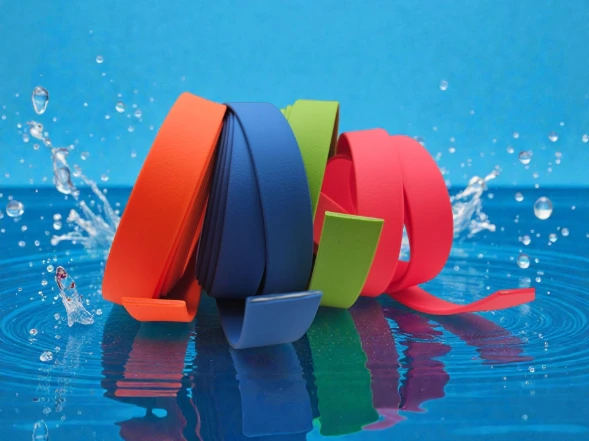Four vibrant rolls of BioThane® material—orange, blue, green, and pink—are displayed on a reflective blue surface with water splashes, highlighting their waterproof, durable, and wipe-clean properties, ready to be handcrafted into custom dog and horse gear.