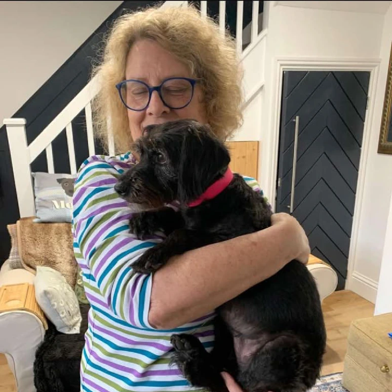 A small black dog is being cuddled indoors by a woman in a striped shirt. The dog is wearing a vibrant pink BioThane® collar, showcasing waterproof, stink-proof, made-to-measure pet gear. The cozy home setting highlights the comfort and joy these handcrafted products bring to pets and their owners.