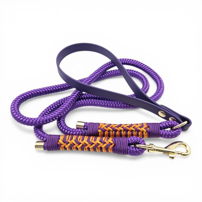 Premium handcrafted dog lead made from vibrant purple BioThane® and rope, featuring an ergonomic handle, gold-tone hardware, and decorative orange and yellow paracord weaving. Fully waterproof, stink-proof, and easy to clean; designed for style, durability, and daily adventures.